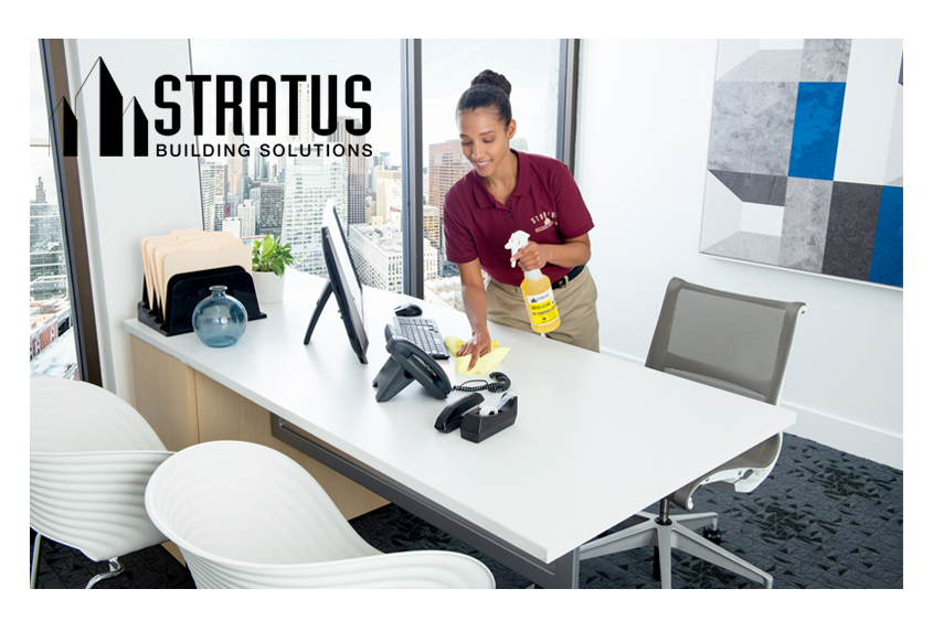 A Smiling Uniformed Stratus Cleaner Holds a Bottle of Cleaning Spray in One Hand While Wiping a Desk with the Other